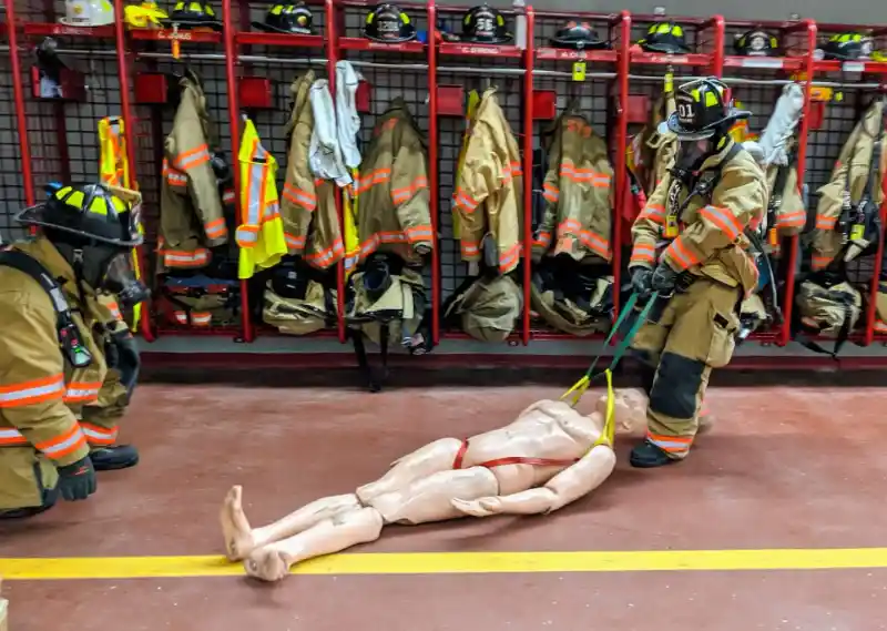 Firefighter in gear, practicing with a dummy in front of the lockers
